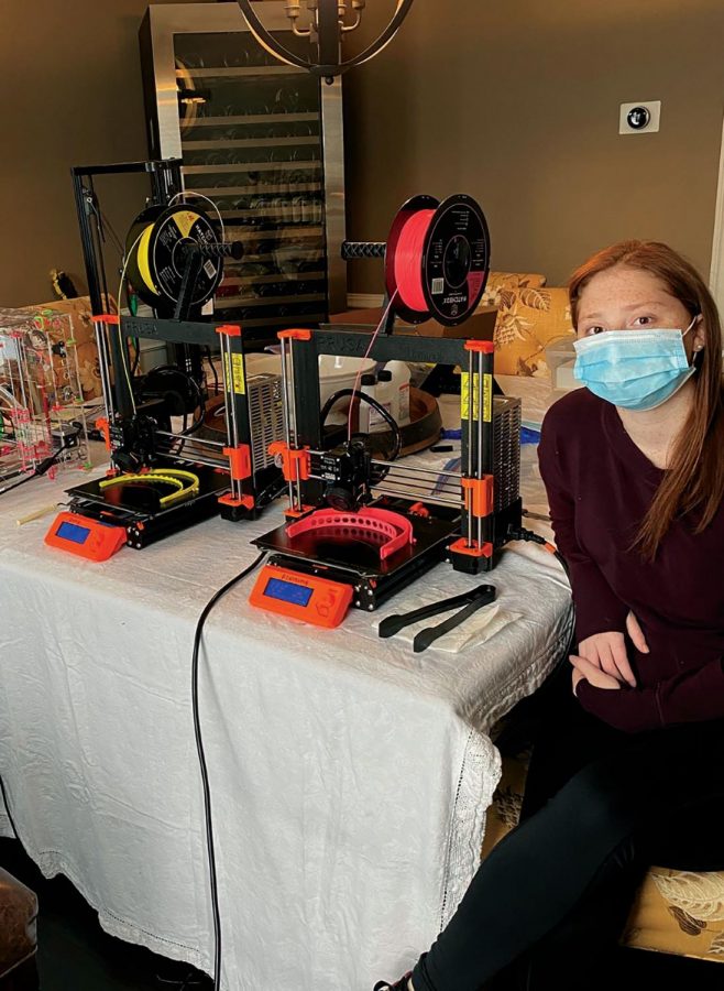 Jenna Bortnick sits next to a Prusa i3 3D-printer as it creates a face shield. 
