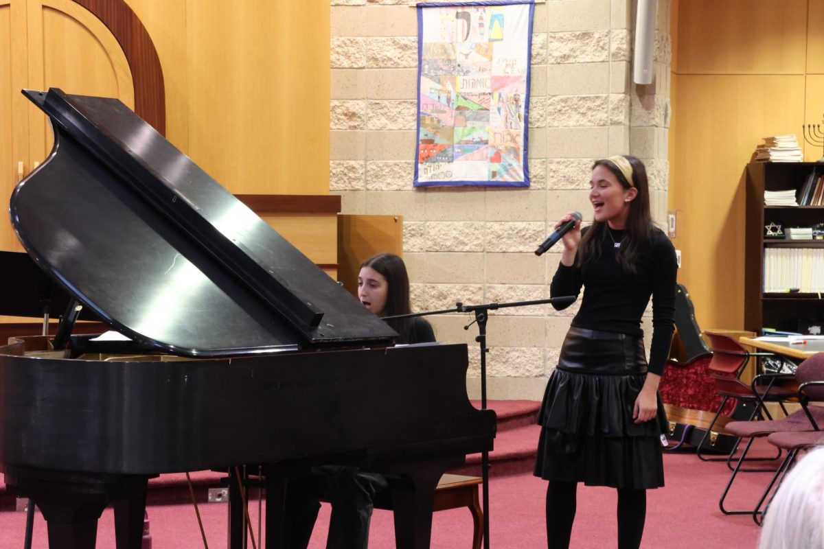 Surrounded by family and friends, juniors Maya Greenblum and Ella Arking perform “Hometown Glory” by Adele in the Feith Beit Midrash. Their performance was one of many small group performances that occurred throughout the night. Students were able to sign up for a five-minute slot and perform whatever they wanted. With parents, students and teachers in the audience, there was a significant turnout in the small group performance section of the event. 

Both Greenblum and Arking have been performing for the school since middle school, at Kab Shabs, spring and winter concerts and Arts Chai Lights. Arking has been a member of the Shir Madness a cappella group and is in Instrumental Ensemble, and Greenblum is currently a member of Shir Madness and a conductor for the group. Arking and Greenblum have performed together at many JDS events, with the event on Wednesday night being their last Arts Chai Lights together.

“Ella and I have been performing together for quite a while,” Greenblum said. “The first time we performed together was middle school, so it was very special to be able to perform with her again at Arts Chai-Lights. We picked a song that we both really loved and have heard a lot together, and we made sure to practice it. I think the piano was beautiful, but overall it was very special to perform a song with Ella, when we have such a deep connection together when it comes to music and singing and performances.”